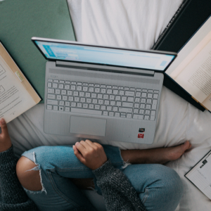 A person sits on a bed working at a laptop surrounded by various books and notebooks